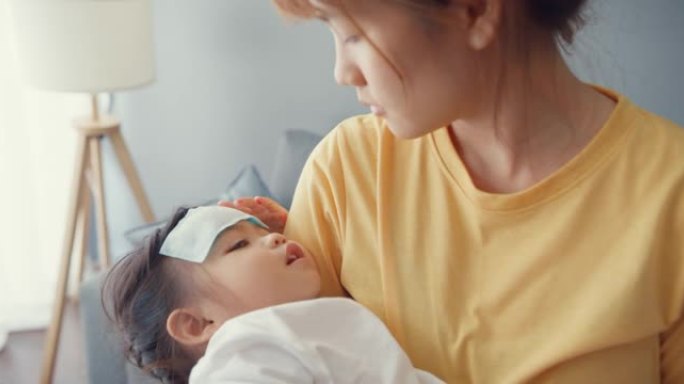 快乐快乐的亚洲家庭妈妈抱着生病的蹒跚学步的女孩，把退热凝胶贴在婴儿额头上，走在家里的客厅。