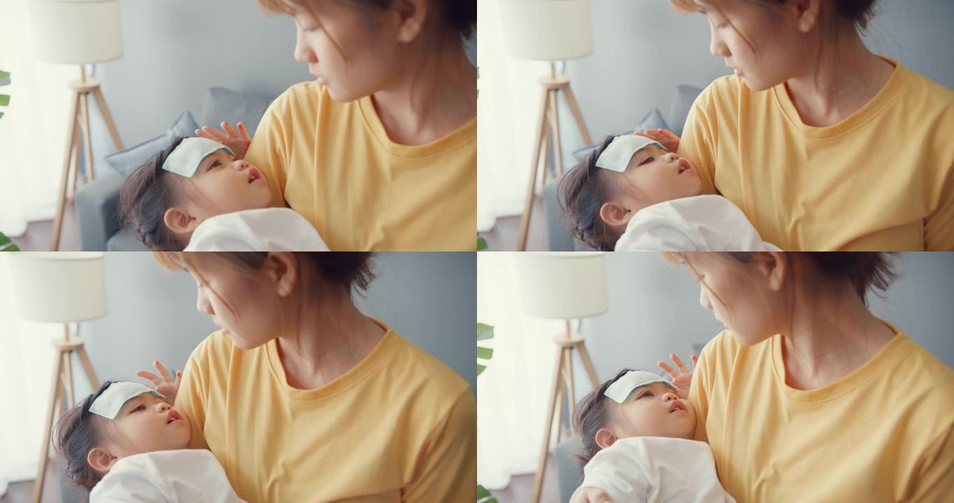 快乐快乐的亚洲家庭妈妈抱着生病的蹒跚学步的女孩，把退热凝胶贴在婴儿额头上，走在家里的客厅。