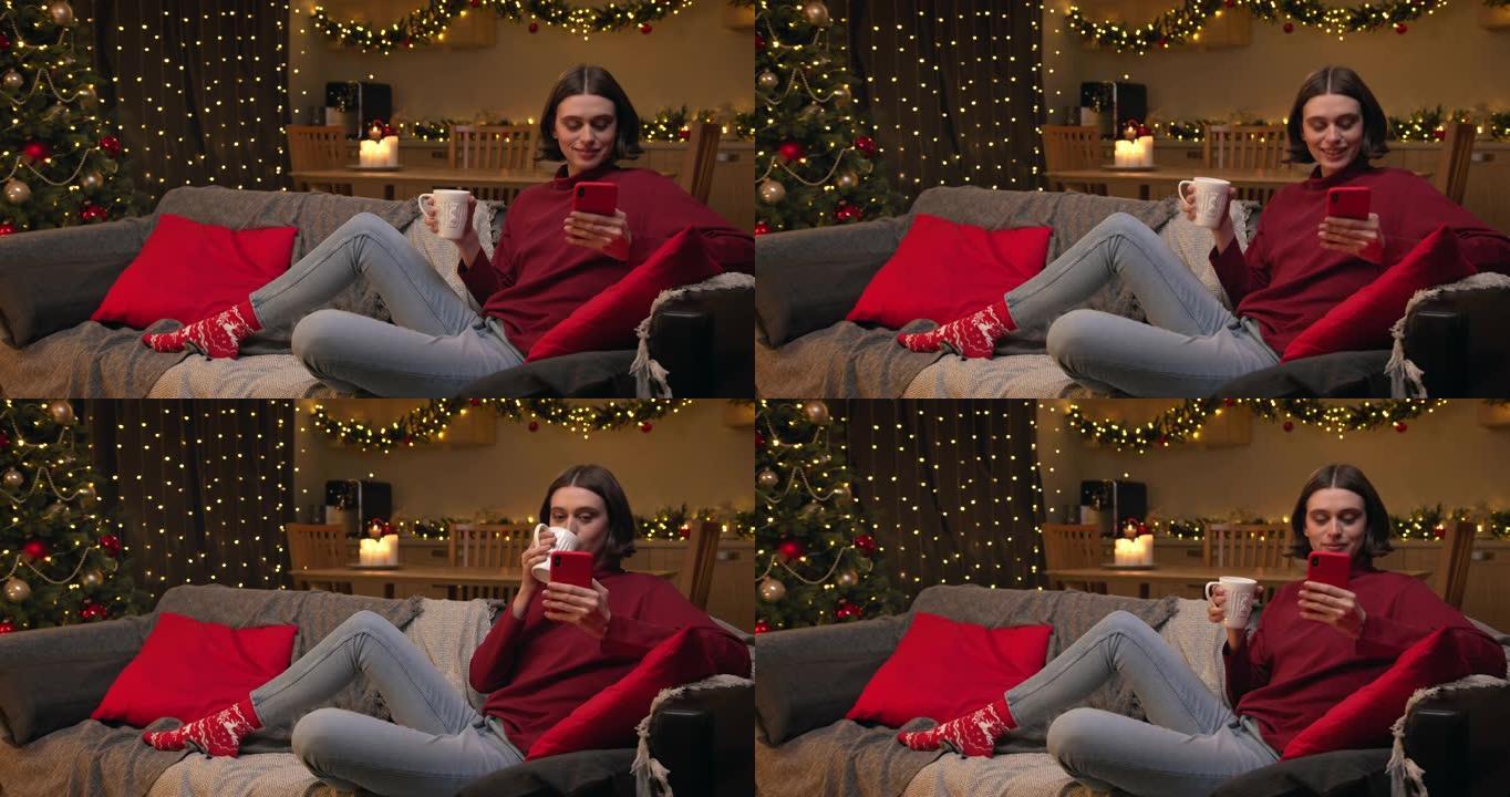 一个穿着红色毛衣和牛仔裤的棕色头发的年轻迷人女人坐在一个舒适的圣诞节装饰的房间里，在她的红色智能手机