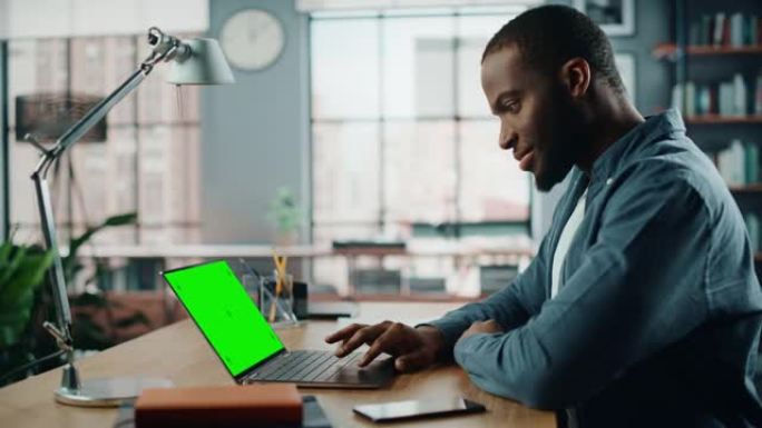 英俊的黑人非洲裔美国专家在家庭客厅的笔记本电脑上工作，绿屏模拟显示。自由职业者在社交网络上通过互联网
