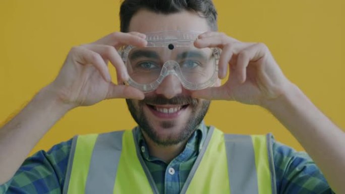 穿着安全背心的年轻人的慢动作肖像穿上工业护目镜，在黄色背景上微笑