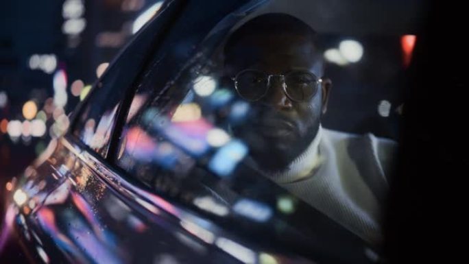 时髦的戴眼镜的黑人在雨夜坐在出租车的后座上通勤回家。英俊的男性乘客在城市街道上开着霓虹灯的汽车时看着