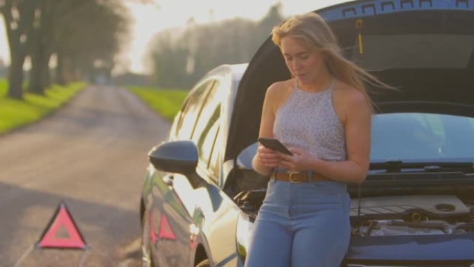 乡间小路上的危险警告标志，一名女子站在抛锚的汽车旁边，引擎盖朝上，用手机发短信寻求帮助-慢动作拍摄