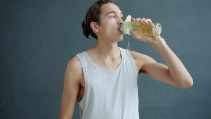 运动员的慢动作肖像从瓶子里喝水，然后看着相机，在灰色背景上微笑