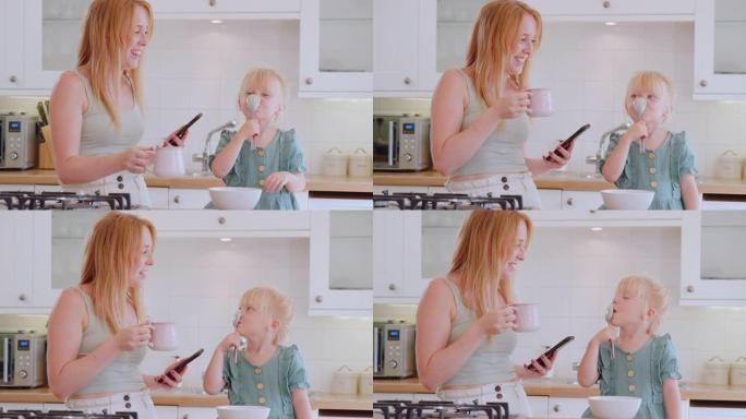 厨房里的女孩试图把勺子挂在鼻子上吃早餐，而母亲使用手机