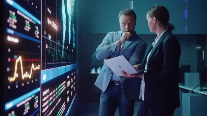 项目经理和计算机科学工程师在使用大屏幕显示器和笔记本电脑时交谈，显示基础设施信息图表数据。电信公司系