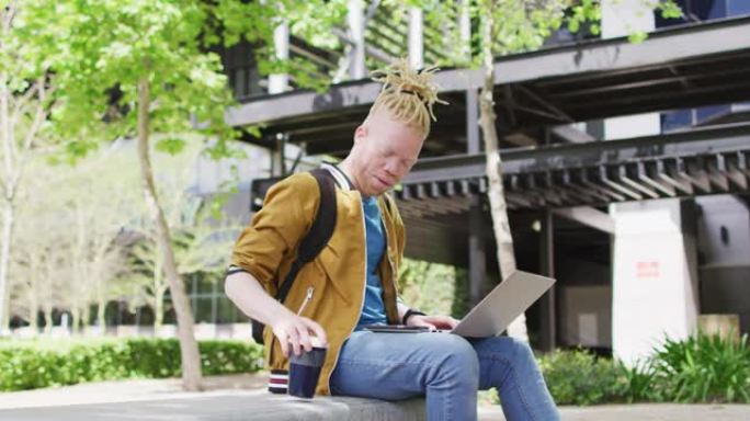 体贴的白化病非裔美国人，留着辫子，坐在公园里喝咖啡，使用笔记本电脑