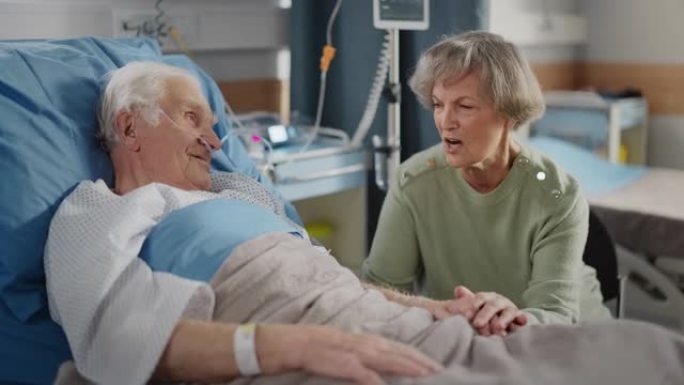 医院病房: 老人躺在床上休息，他有爱心的美丽妻子来访，支持他坐在旁边，手牵着手，幸福的夫妻在一起聊天