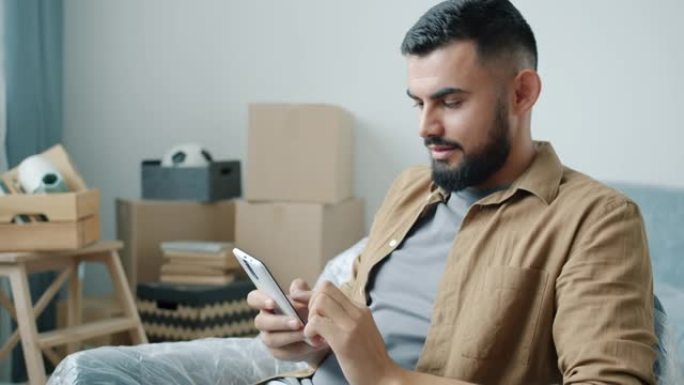 快乐的中东男子使用智能手机触摸屏坐在带纸板箱的新公寓里