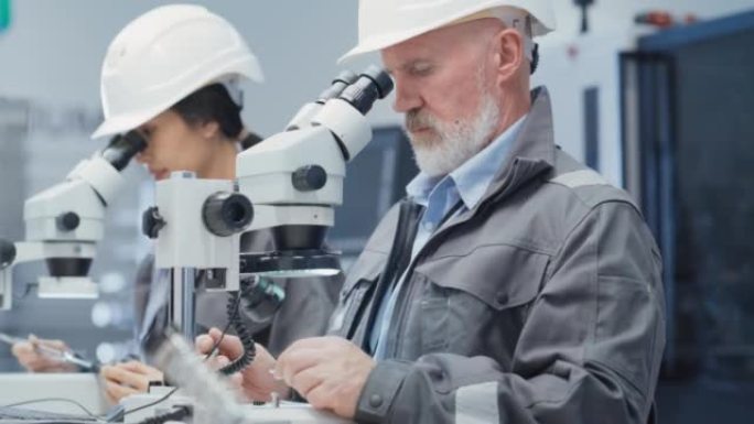 中年工业科学家和年轻的亚洲工程师在工厂的办公桌上工作，使用显微镜处理小型制造生产零件和细节。
