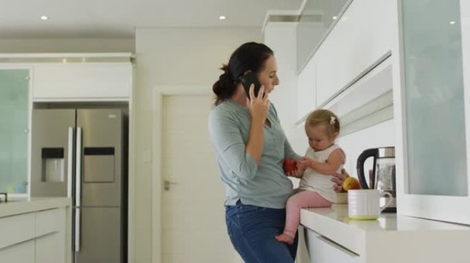 高加索母亲抱着婴儿在家里的厨房里用智能手机聊天