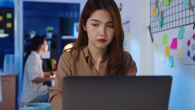 快乐亚洲女商务人士在新常态下保持社交距离以防病毒，同时使用笔记本电脑在线商务，加班回到办公室晚上工作