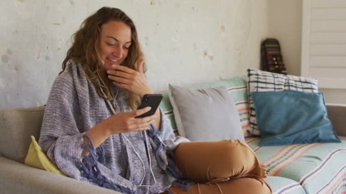 微笑的白人妇女穿着毯子，坐在沙发上，使用智能手机在小屋客厅