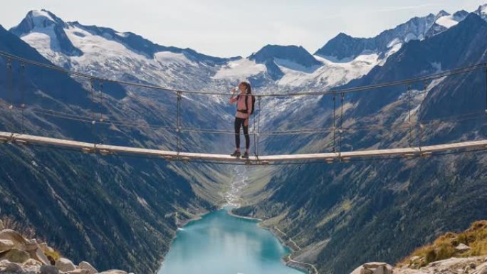 女徒步旅行者休息一下，站在山涧上的吊桥上，欣赏山谷湖和周围群山的壮丽景色