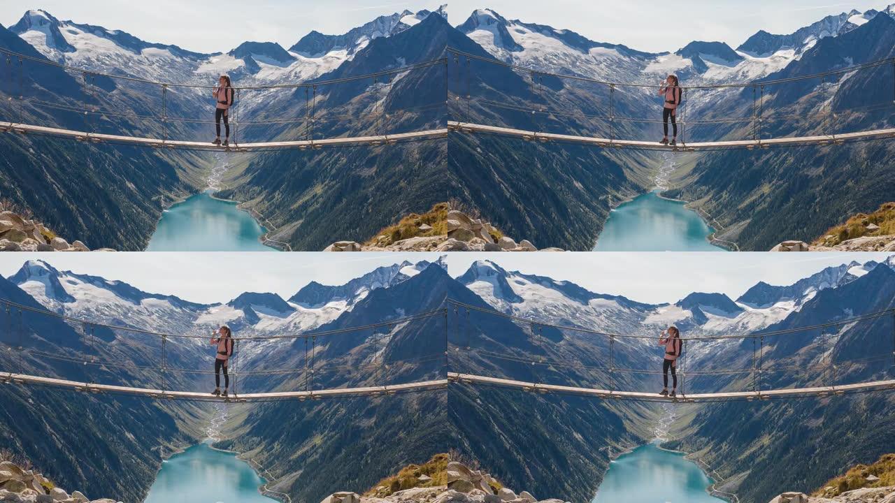 女徒步旅行者休息一下，站在山涧上的吊桥上，欣赏山谷湖和周围群山的壮丽景色
