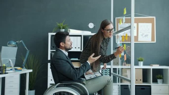 坐在轮椅上的年轻女子和残疾男子在办公室玻璃板上的便签上聊天
