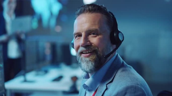 大胡子快乐的高级技术支持专家在计算机上工作时正在耳机上交谈。成功的员工转向镜头，轻轻地微笑。带显示屏