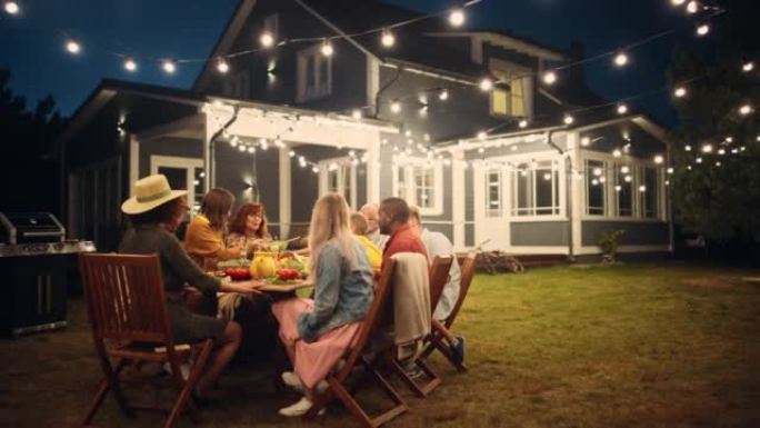 父母，孩子和朋友聚集在一个带有灯光装饰的美丽家外的烧烤餐桌旁。老人和年轻人玩得开心，吃东西。后院的花