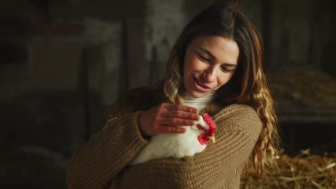 年轻快乐的女农场主的电影镜头在乡村农业农场的干草仓里爱抚着生态生长的白母鸡，用于生物真正的食品行业。