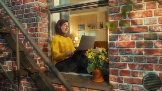 年轻的穆斯林女性坐在舒适的褐砂石房屋公寓的窗台上，在笔记本电脑上进行视频通话。快乐女孩与朋友、家人或