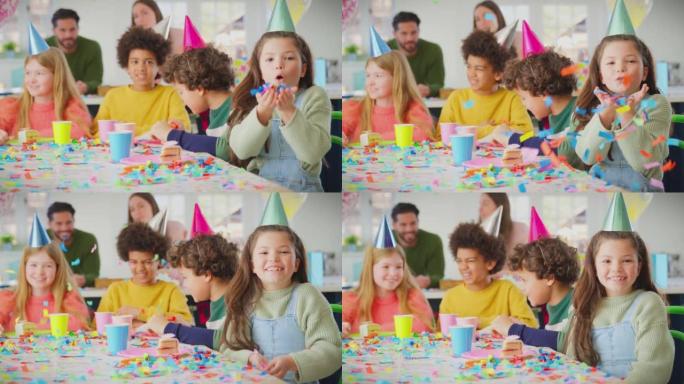 在生日聚会上与朋友和父母在家的女孩在相机上吹五彩纸屑