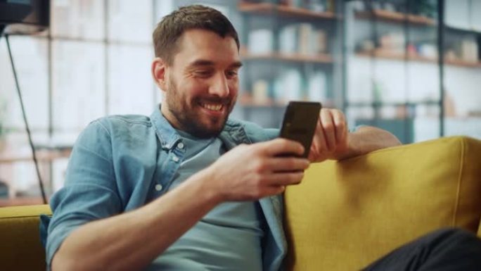 快乐英俊的白人男人在家里舒适的客厅使用智能手机。男人躺在舒适的沙发上。他正在浏览互联网，并在社交网络