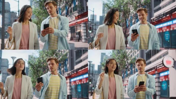 年轻时尚的多民族夫妇在大城市的街道上随意行走。英俊的白人男性向美丽的日本女性展示智能手机。不同的朋友