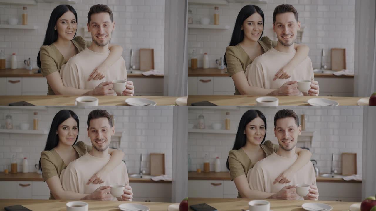 丈夫和妻子一起站在厨房里的慢动作肖像，拿着咖啡杯，微笑着