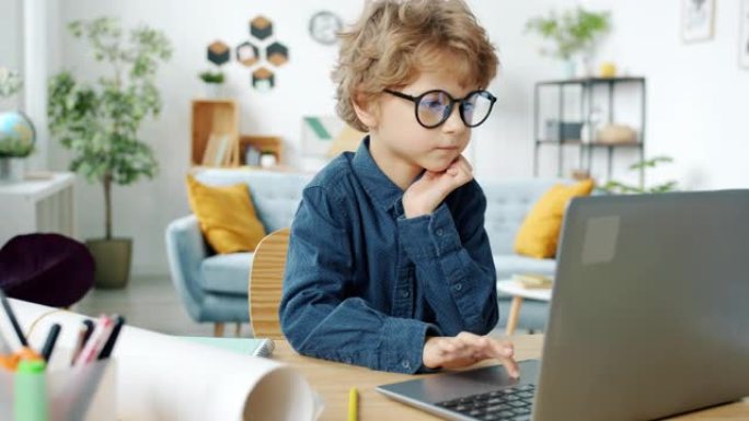 戴眼镜的严肃孩子坐在家里的桌子上，用笔记本电脑工作
