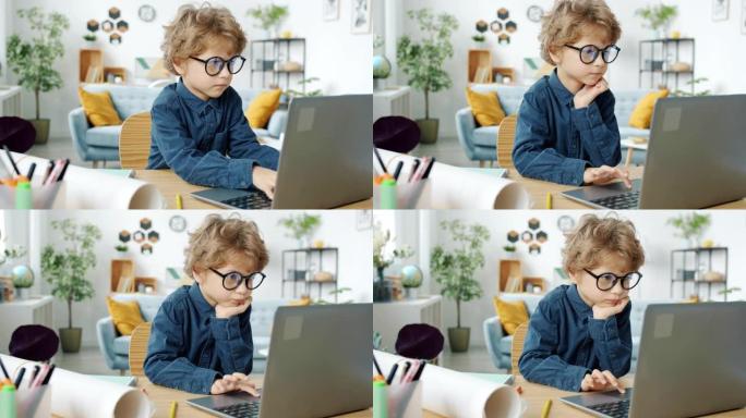 戴眼镜的严肃孩子坐在家里的桌子上，用笔记本电脑工作
