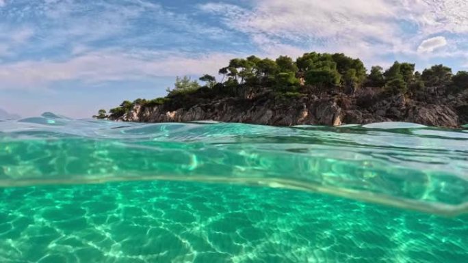 半水下慢动作观看沙滩底部和树木岩石。希腊哈尔基迪基西托尼亚的清澈海水。用圆顶从空气和水的边界拍摄