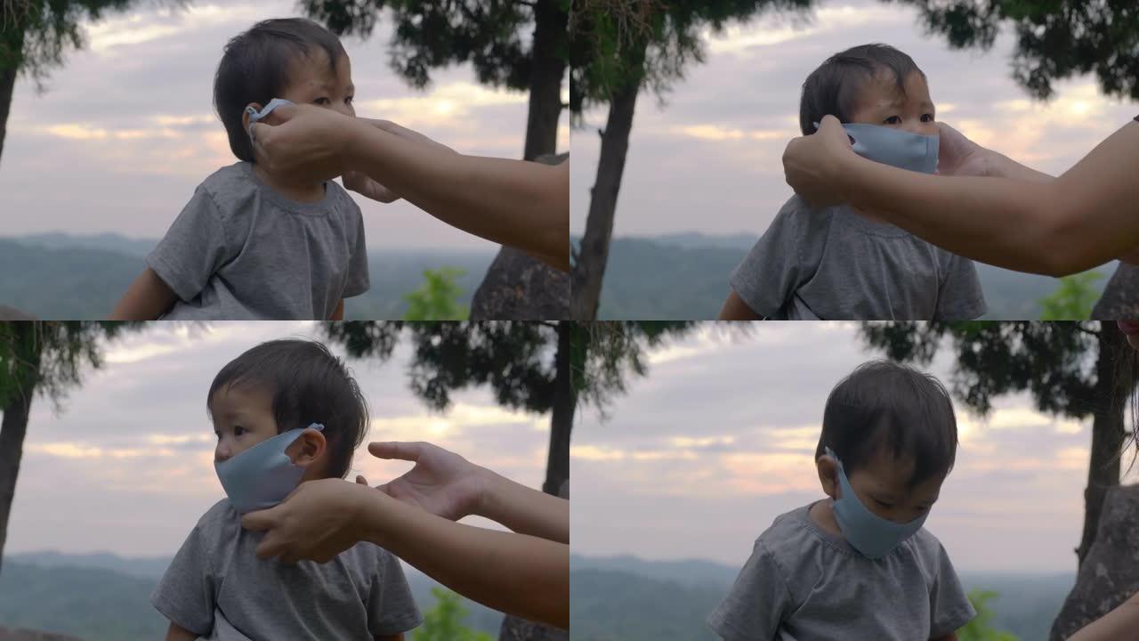 亚洲母亲为14个月大的儿子戴上医用口罩，以预防冠状病毒或Covid-19的流行，在日落时分一起户外活