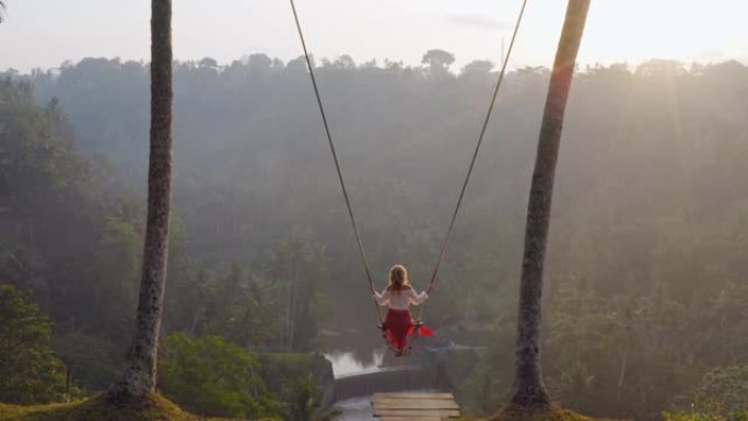 鸟瞰图女人在日出时在热带雨林上摇摆，坐在秋千上，欣赏河流的乐趣，享受假日旅行自由