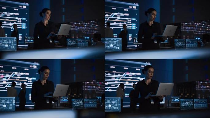 美丽自信的女项目经理站在电信系统控制室的笔记本电脑上工作。在后台大屏幕中显示基础设施、图表、系统分析