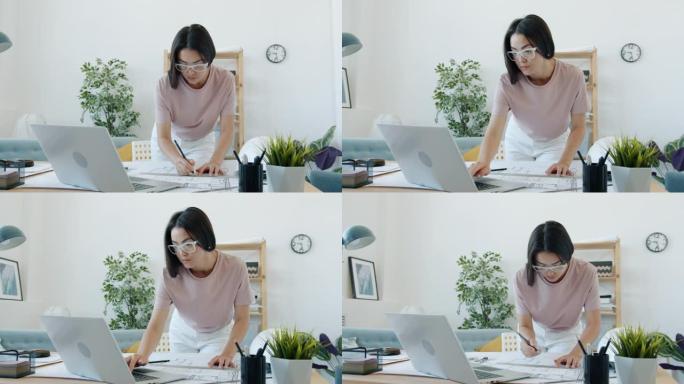 年轻的女建筑师在建筑平面图上绘制蓝图并在家中使用笔记本电脑