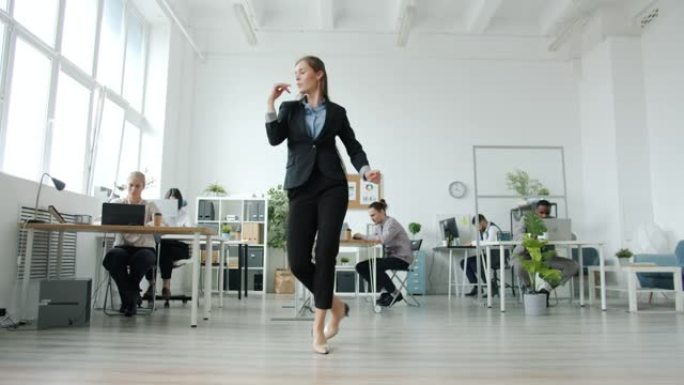 优雅的年轻女子在工作场所跳舞，而员工在办公桌前工作，则转向音乐