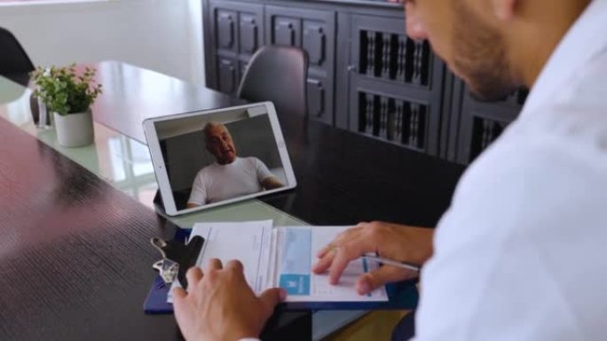 男医生在家中使用平板电脑进行视频通话，并与一名高级患者描述症状，同时在病历上做笔记