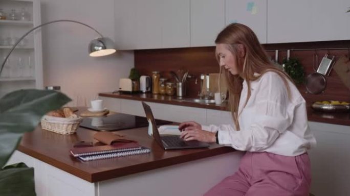 美丽轻松的高加索女商人使用笔记本电脑在豪华家庭厨房餐桌上在线工作。工作狂的概念。