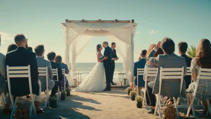 美丽的新娘和新郎在海边的海滩上举行户外婚礼。浪漫情侣结婚、接吻和拥有多民族文化的朋友庆祝的完美场所。