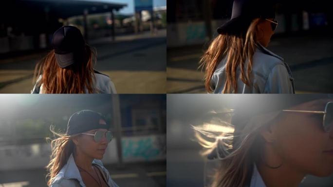 日落时分，棕色头发的特写年轻女子戴着牛仔夹克太阳镜，戴着帽子走在海滩旁边