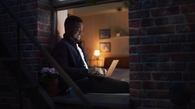 英俊，面带微笑的非洲黑人男子坐在家里的窗台上在笔记本电脑上工作。一位年轻的男性自由职业者在计算机上工