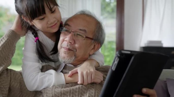 在家庭中的亲密关系中，与亚洲老前辈祖父母在家中与计算机平板电脑上的技术一起玩耍和观看。年轻女孩从后面