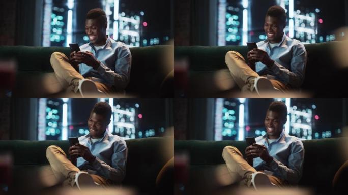 兴奋的黑人非裔美国人坐在客厅的沙发上使用智能手机。快乐的男人在家里微笑，通过互联网与朋友聊天。使用社