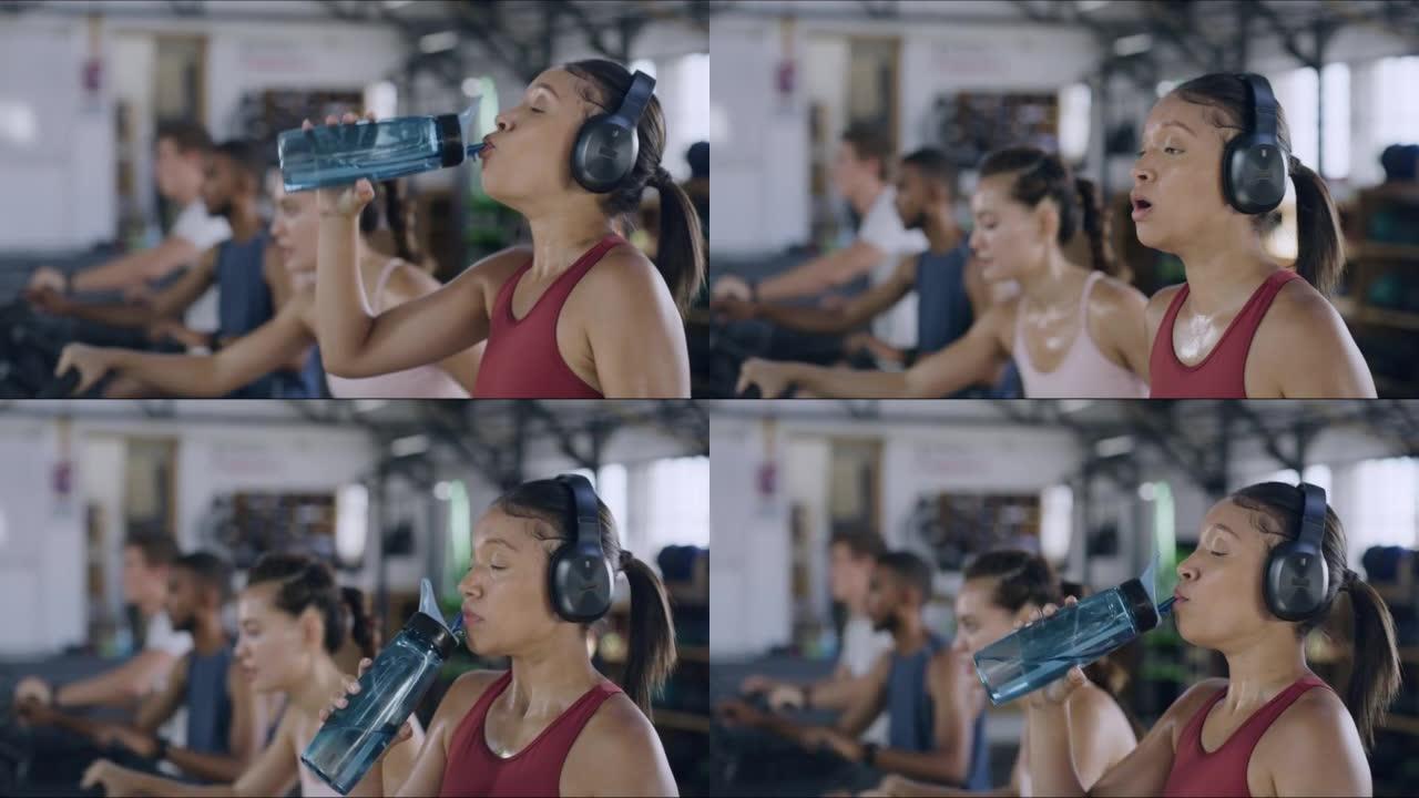 出汗的女运动员在锻炼期间在繁忙的健身房里喝水。疲倦和口渴的运动女性在锻炼时补水和提神。活跃的健身女士