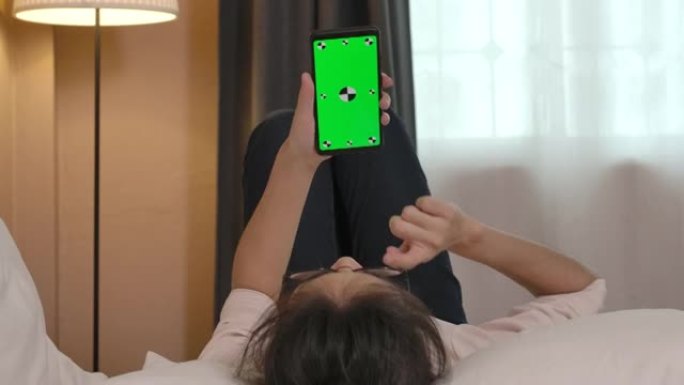 年轻的亚洲妇女躺在床上，使用智能手机与绿屏chromakey上的人进行视频通话。检疫期间，一个女孩在