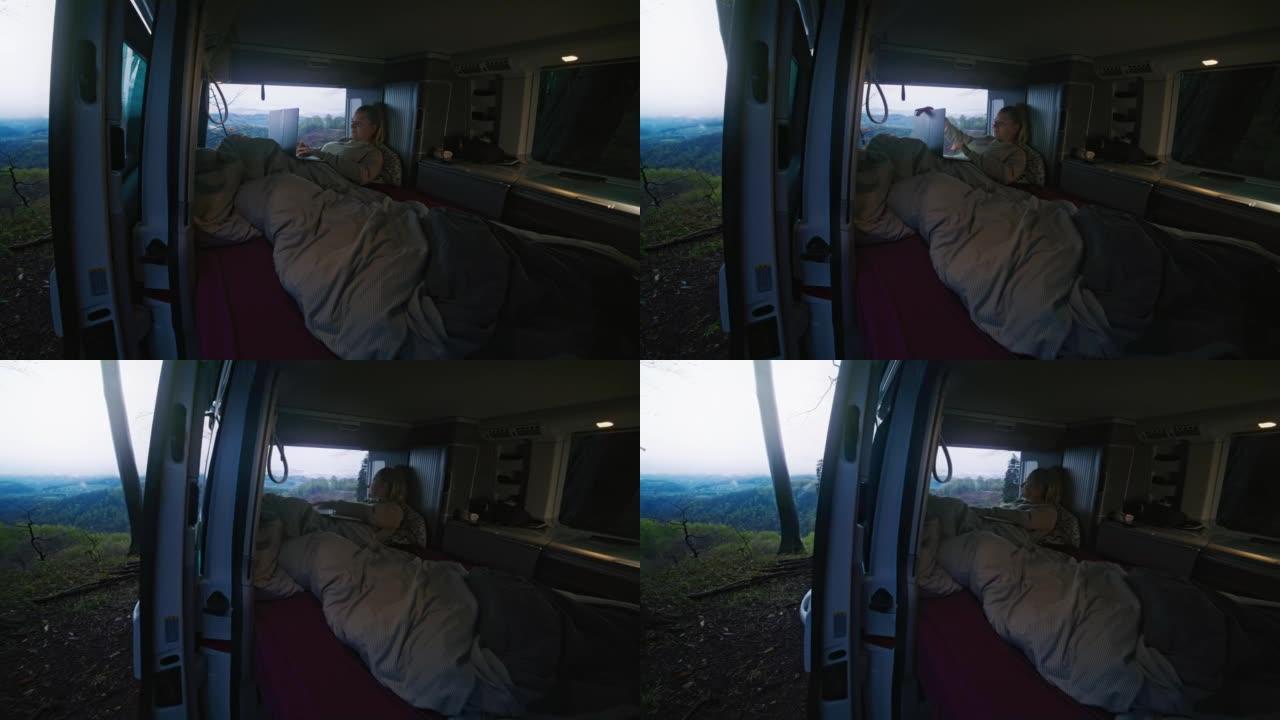金发碧眼的女人躺在毯子里躺在面包车里时，停止在笔记本电脑上工作，看着风景，从户外看，面包车的门打开了