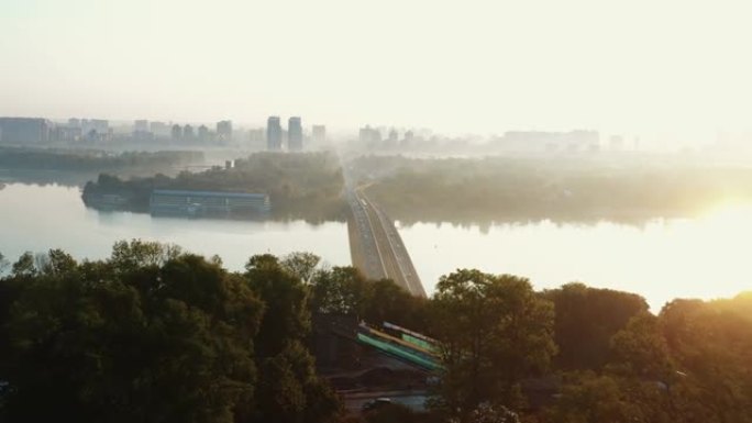 无人机飞过夏季的树木，展示了令人惊叹的汽车桥，第聂伯河上的火车，田园般的夏季日出基辅。