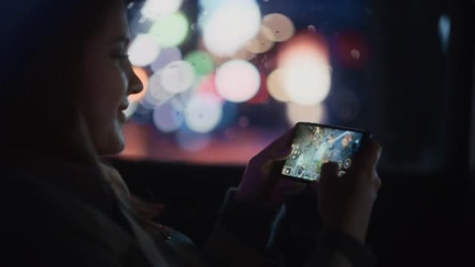 快乐的女性晚上坐出租车后座回家。美丽的女人乘客在智能手机上玩策略视频游戏，而在城市街道上的一辆带有工