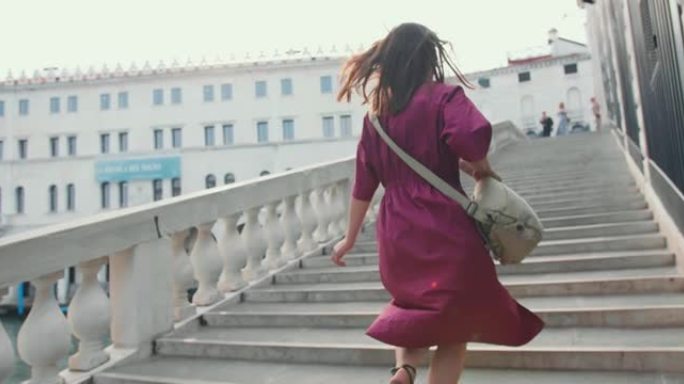 后视年轻快乐的黑发白人旅游妇女，穿着紫色连衣裙走上威尼斯的旧城大桥楼梯。