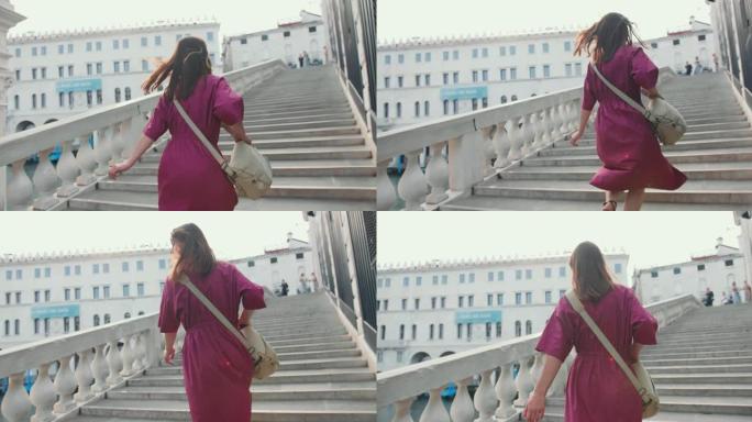 后视年轻快乐的黑发白人旅游妇女，穿着紫色连衣裙走上威尼斯的旧城大桥楼梯。